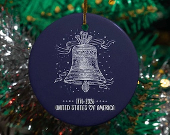 May include: A round, navy blue Christmas ornament with a white illustration of the Liberty Bell. The ornament has the text "1776-2026 UNITED STATES OF AMERICA" in white. A gold string is attached for hanging.