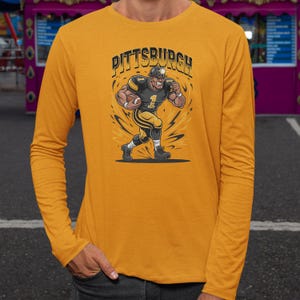 May include: Mustard yellow long-sleeve shirt featuring a cartoon illustration of a football player in black and gold uniform. The word "Pittsburgh" is printed above the player. The player is running with a football.