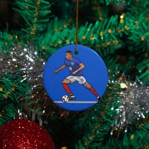 May include: A blue ceramic Christmas ornament featuring a soccer player in a blue jersey with the number 10, white shorts, and red socks. The player is kicking a soccer ball. The ornament is round and has a gold string for hanging.