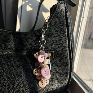 May include: Black leather handbag with a silver zipper and strap. A decorative keychain with pink, purple, and beige buttons hangs from the strap. The buttons are various sizes and have four holes. The bag is in front of a window.