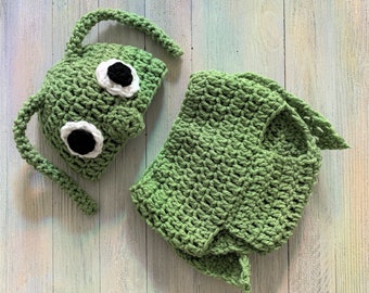 Könnte beinhalten: Grün gehäkelte Baby-Mütze und passendes Outfit mit großen weißen und schwarzen Augen.