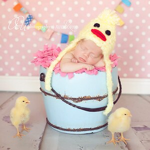 K&ouml;nnte beinhalten: Ein neugeborenes Baby tr&auml;gt eine gelbe, geh&auml;kelte Entenm&uuml;tze und schl&auml;ft in einem blauen Eimer mit rosa Stoff. Zwei gelbe K&uuml;ken sind auf dem Boden.