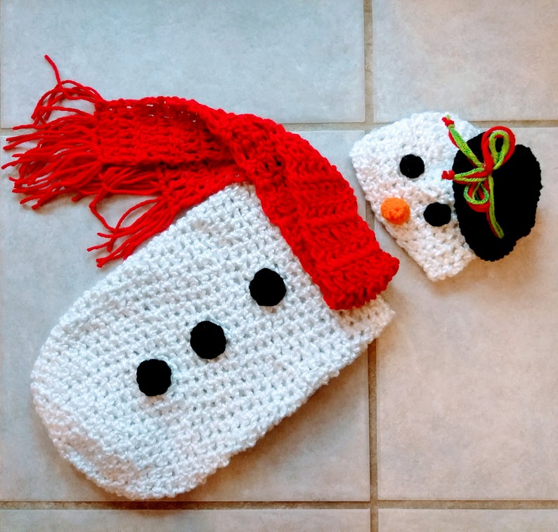 May include: White crocheted snowman outfit with a red scarf and a black and white crocheted hat. The hat has a green and white trim.