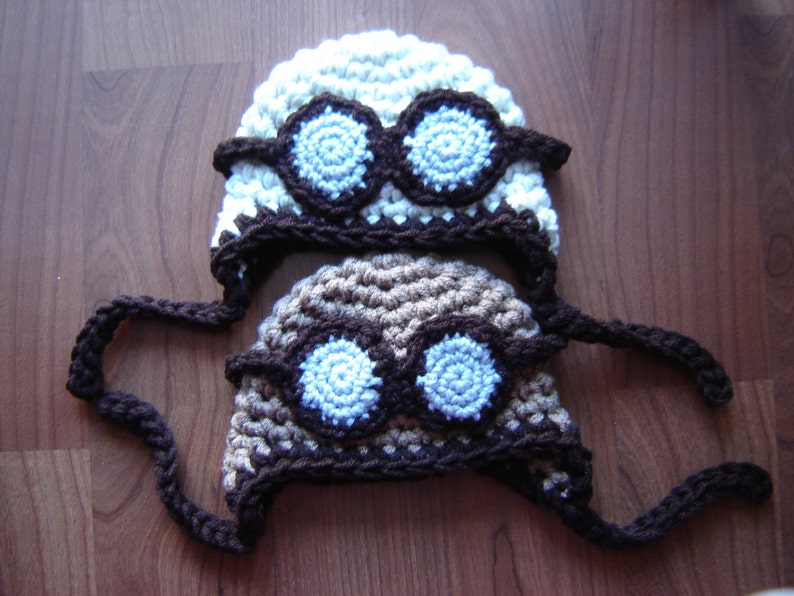 May include: Two crocheted hats with goggles. One hat is white with brown trim and the other is brown with white trim. Both hats have blue goggles.