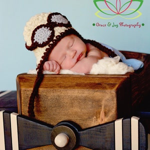 May include: A newborn baby wearing a brown and beige crocheted aviator hat with goggles sleeps in a wooden airplane prop. The airplane prop is painted black with white stripes. The background is a light blue.