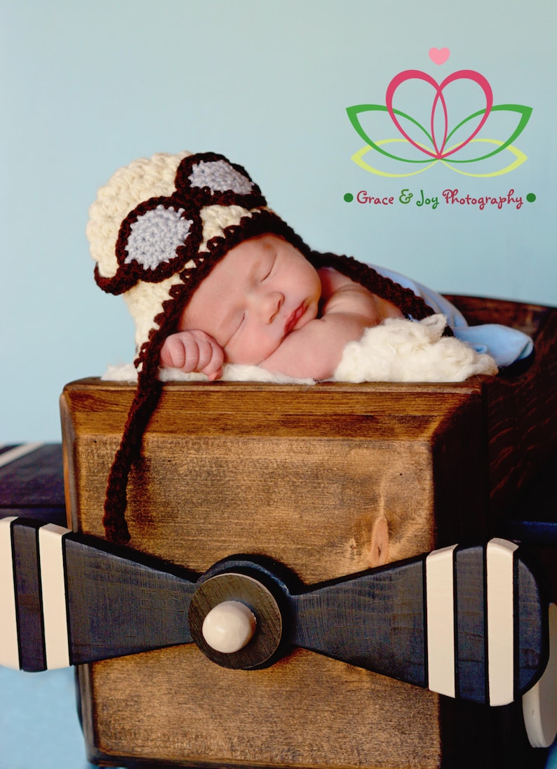 May include: A newborn baby wearing a brown and beige crocheted aviator hat with goggles sleeps in a wooden airplane prop. The airplane prop is painted black with white stripes. The background is a light blue.