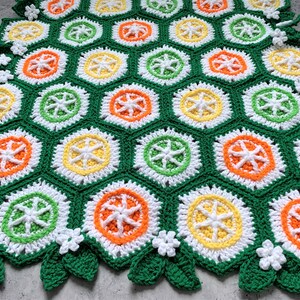 May include: A crocheted blanket with a repeating pattern of orange, yellow, green, and white hexagons. The blanket has a border of green leaves and white flowers.