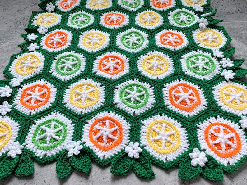 May include: A crocheted blanket with a repeating pattern of orange, yellow, green, and white hexagons. The blanket has a border of green leaves and white flowers.