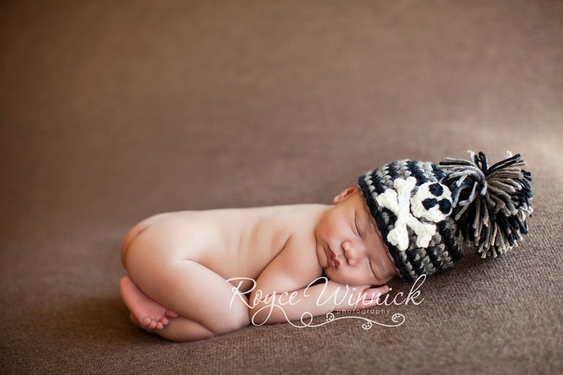 May include: A newborn baby wearing a black, white, and gray crocheted hat with a skull and crossbones design. The hat has a fluffy pom-pom on top.
