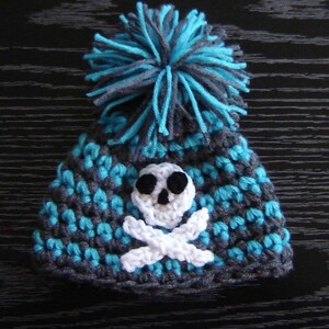 May include: A gray and turquoise crocheted hat with a white skull and crossbones design and a turquoise and gray pom-pom on top.