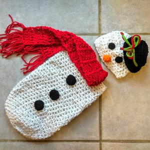 May include: White crocheted snowman outfit with a red crocheted scarf and a white crocheted hat with black and green details.