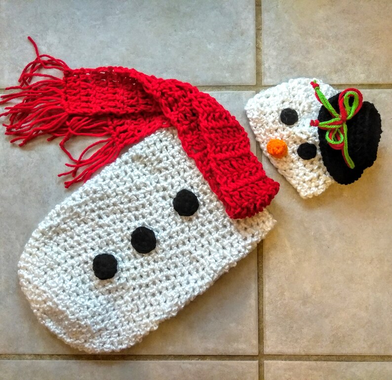 May include: White crocheted snowman outfit with a red crocheted scarf and a white crocheted hat with black and green details.