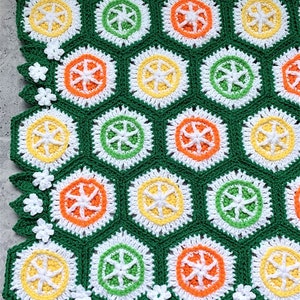 May include: A green crochet blanket with a repeating pattern of hexagons featuring orange, yellow, and green circles. The blanket has a white crocheted border with green leaves and white flowers.