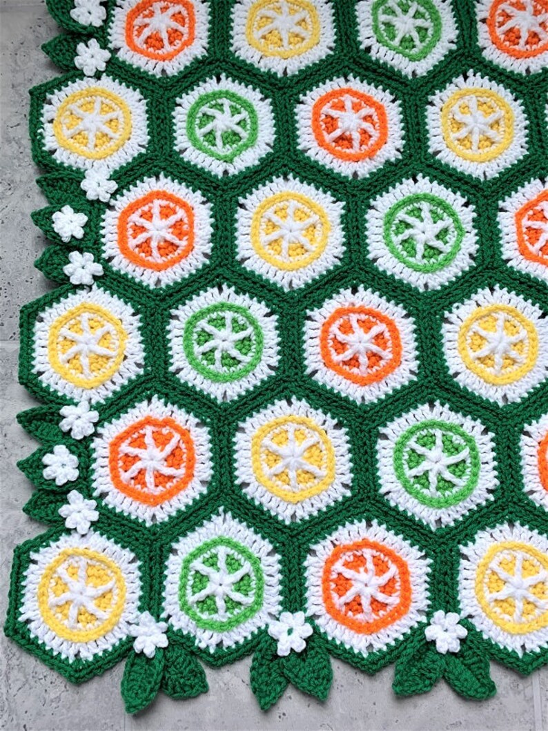 May include: A green crochet blanket with a repeating pattern of hexagons featuring orange, yellow, and green circles. The blanket has a white crocheted border with green leaves and white flowers.