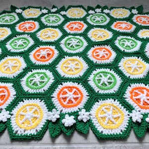 May include: A crocheted blanket with a hexagonal pattern in shades of green, white, yellow, and orange. The blanket features a repeating design of stylized citrus fruits.
