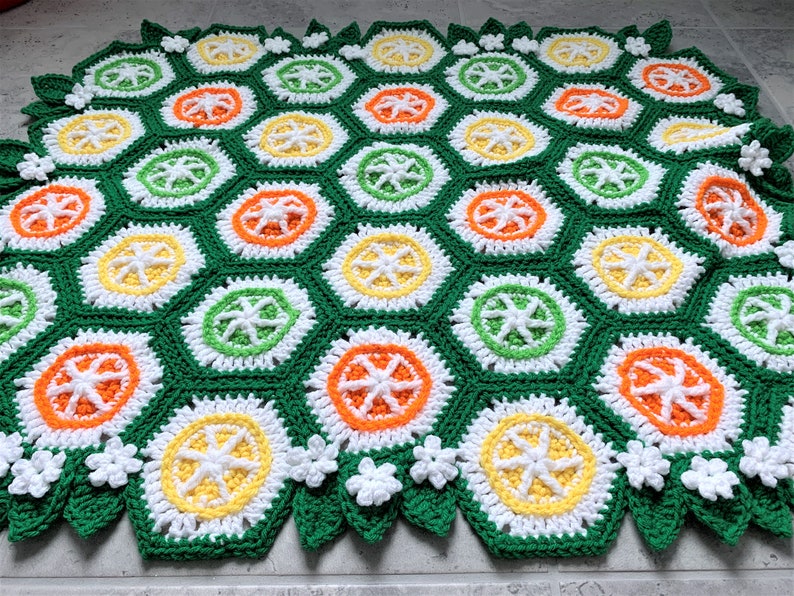 May include: A crocheted blanket with a hexagonal pattern in shades of green, white, yellow, and orange. The blanket features a repeating design of stylized citrus fruits.