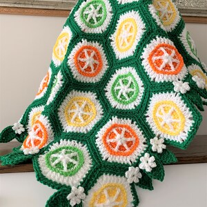 May include: A green, white, orange, and yellow crocheted blanket with a citrus fruit design. The blanket has a scalloped edge with crocheted flowers and leaves.