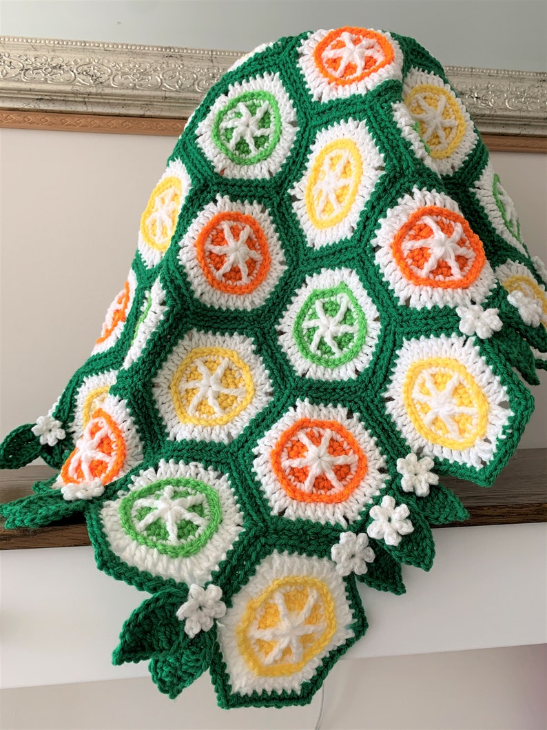 May include: A green, white, orange, and yellow crocheted blanket with a citrus fruit design. The blanket has a scalloped edge with crocheted flowers and leaves.