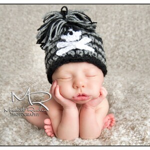 May include: A newborn baby wearing a black and white crocheted hat with a skull and crossbones design. The baby is sleeping with their hands tucked under their chin.