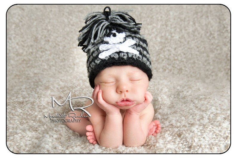 May include: A newborn baby wearing a black and white crocheted hat with a skull and crossbones design. The baby is sleeping with their hands tucked under their chin.
