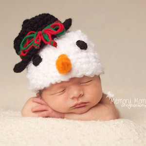 Puede incluir: Un beb&eacute; durmiendo con un gorro de mu&ntilde;eco de nieve blanco y negro con un lazo rojo y verde. El gorro tiene ojos negros y una nariz naranja brillante.