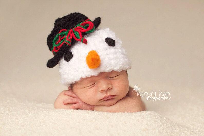 Puede incluir: Un beb&eacute; durmiendo con un gorro de mu&ntilde;eco de nieve blanco y negro con un lazo rojo y verde. El gorro tiene ojos negros y una nariz naranja brillante.