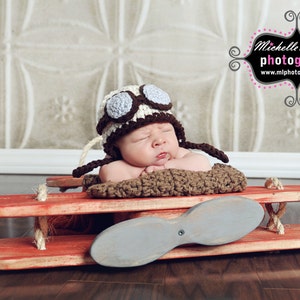 May include: A baby wearing a brown and white crocheted aviator hat with a blue and white propeller on a red wooden toy airplane.