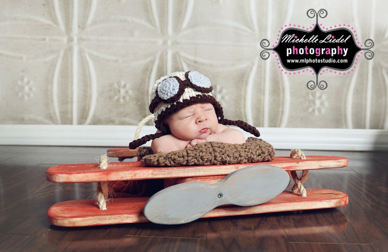 May include: A baby wearing a brown and white crocheted aviator hat with a blue and white propeller on a red wooden toy airplane.