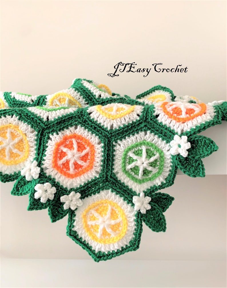 May include: A crocheted blanket with a green background and a pattern of orange, yellow, and white citrus fruit slices. The blanket has a border of green leaves and white flowers.
