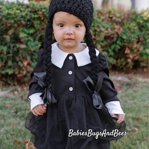 May include: A baby dressed in a black dress with a white collar and cuffs, styled to resemble a character. The outfit includes a black crocheted hat and long, braided pigtails. The text "BabiesBugsAndBees" is visible at the bottom.