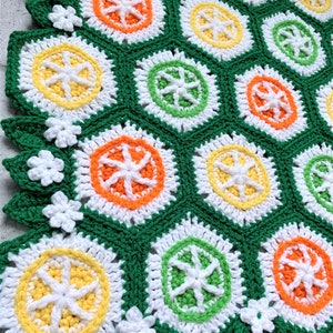 May include: A crocheted blanket with a repeating pattern of green, white, orange, and yellow circles. The blanket has a scalloped edge with white flowers.