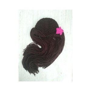 May include: A brown crochet hat with long, dark brown yarn strands resembling hair. A small, pink flower is attached to the side of the hat. This handmade item is likely a costume piece or a photo prop.
