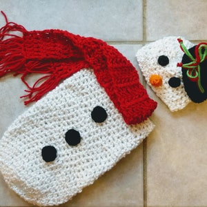 May include: White crocheted snowman baby outfit with a red scarf and a matching white crocheted hat with black eyes, a carrot nose, and a black and green hat.