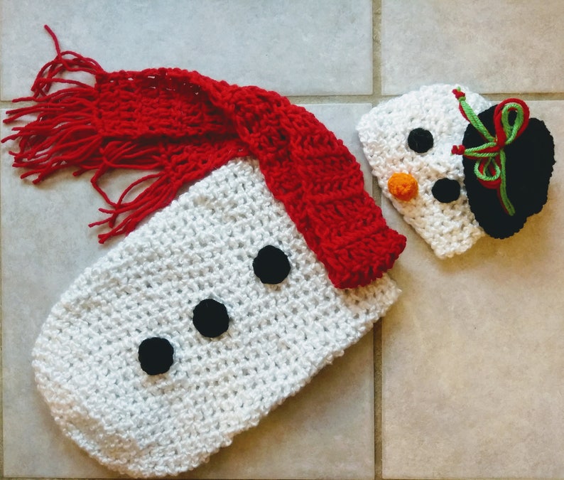 May include: White crocheted snowman baby outfit with a red scarf and a matching white crocheted hat with black eyes, a carrot nose, and a black and green hat.
