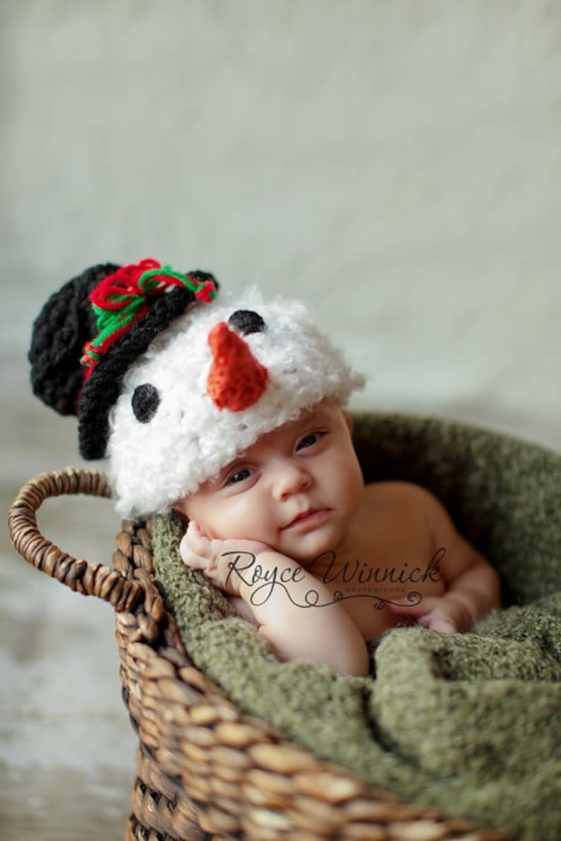 Puede incluir: Un beb&eacute; con un gorro de punto blanco de mu&ntilde;eco de nieve con un sombrero de copa negro, ribetes rojos y verdes y una nariz de zanahoria. El beb&eacute; est&aacute; acostado en una cesta de mimbre marr&oacute;n forrada con tela verde.