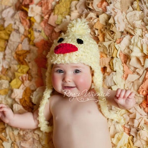 K&ouml;nnte beinhalten: Ein Baby tr&auml;gt eine gelbe, geh&auml;kelte H&uuml;hnerm&uuml;tze mit rotem Schnabel und schwarzen Augen. Das Baby liegt auf einem Bett aus bunten Stoffresten.