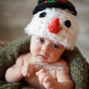 Puede incluir: Un beb&eacute; con un gorro de punto blanco de mu&ntilde;eco de nieve con un sombrero de copa negro, nariz roja y ojos negros. El beb&eacute; est&aacute; sentado en una manta de punto verde.