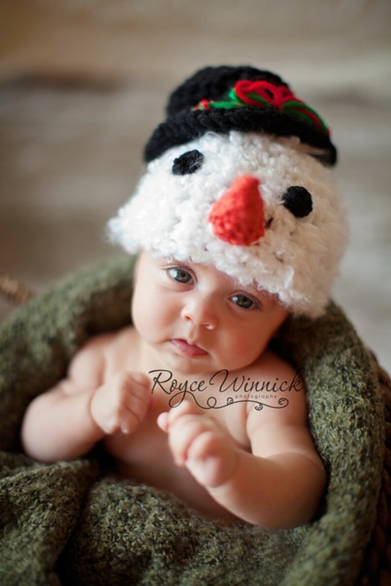 Puede incluir: Un beb&eacute; con un gorro de punto blanco de mu&ntilde;eco de nieve con un sombrero de copa negro, nariz roja y ojos negros. El beb&eacute; est&aacute; sentado en una manta de punto verde.