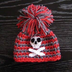 May include: Red and gray crocheted hat with a white skull and crossbones design and a gray and red pom-pom on top.