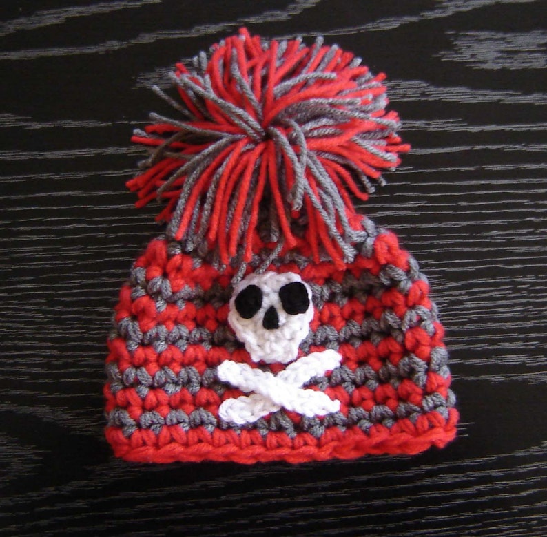 May include: Red and gray crocheted hat with a white skull and crossbones design and a gray and red pom-pom on top.
