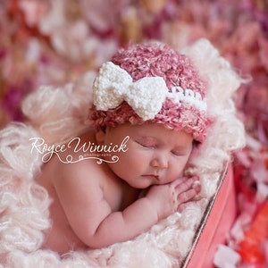 May include: A sleeping baby wearing a pink crocheted hat with a white bow on top, lying on a white fluffy blanket in a pink wooden box. The background is a pink and white flower petal backdrop.