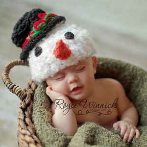 Puede incluir: Un beb&eacute; reci&eacute;n nacido con un gorro de mu&ntilde;eco de nieve de crochet blanco con un sombrero de copa negro, nariz roja y ojos negros, durmiendo en una cesta de mimbre marr&oacute;n forrada con una manta verde. El texto "Royce Winnick Photography" es visible en la imagen.
