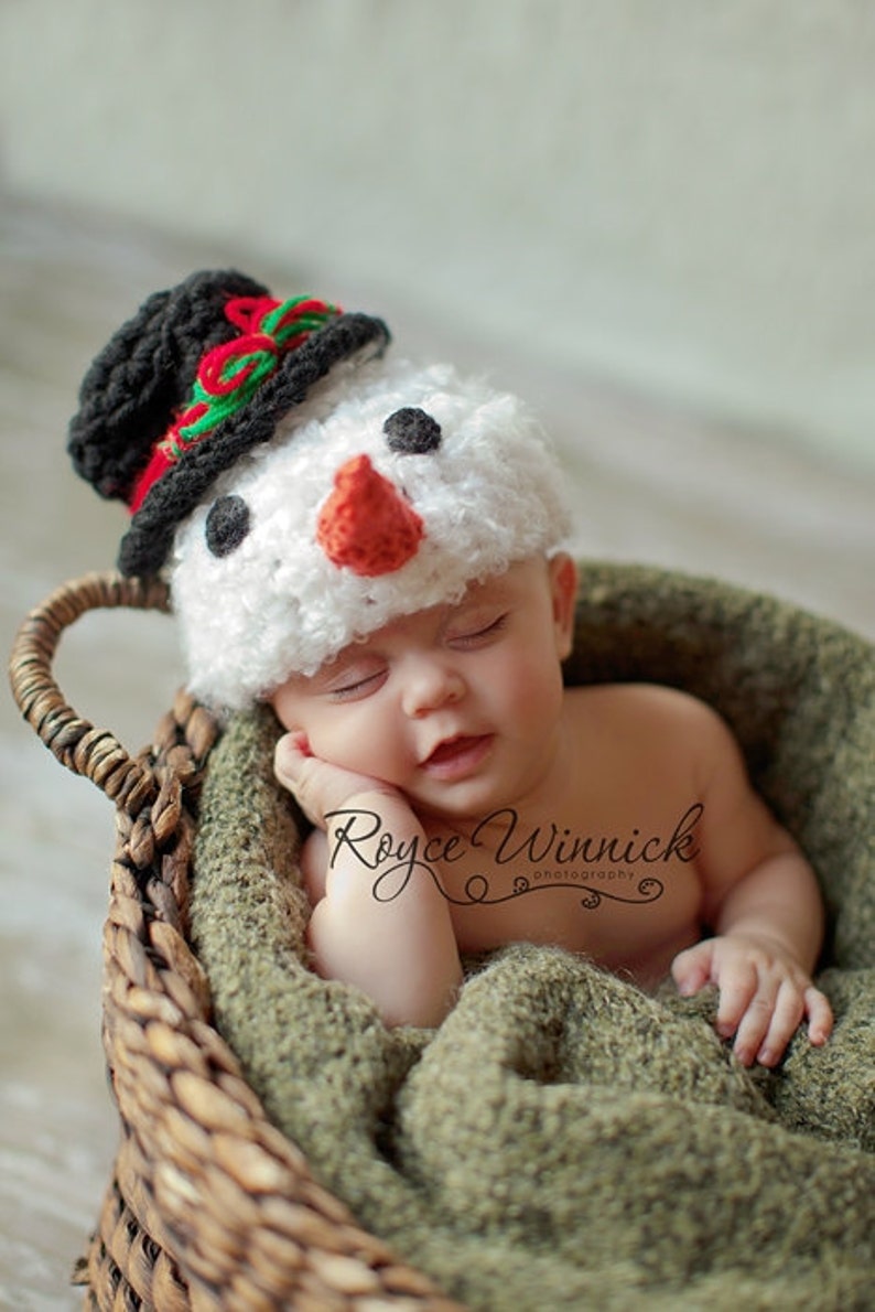 Puede incluir: Un beb&eacute; reci&eacute;n nacido con un gorro de mu&ntilde;eco de nieve de crochet blanco con un sombrero de copa negro, nariz roja y ojos negros, durmiendo en una cesta de mimbre marr&oacute;n forrada con una manta verde. El texto "Royce Winnick Photography" es visible en la imagen.