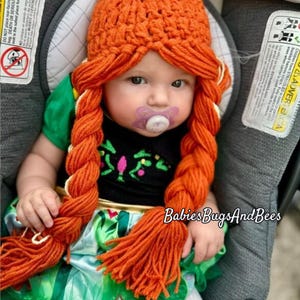 May include: A baby wearing an orange crochet hat with long, braided yarn hair. The baby is also wearing a black top with a green and blue dress. The baby has a pacifier in their mouth. The text "Babies Bugs And Bees" is visible.