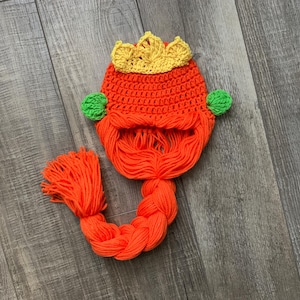 May include: A crocheted hat with a crown, orange hair, and green side pieces. The hat has a long orange braid.