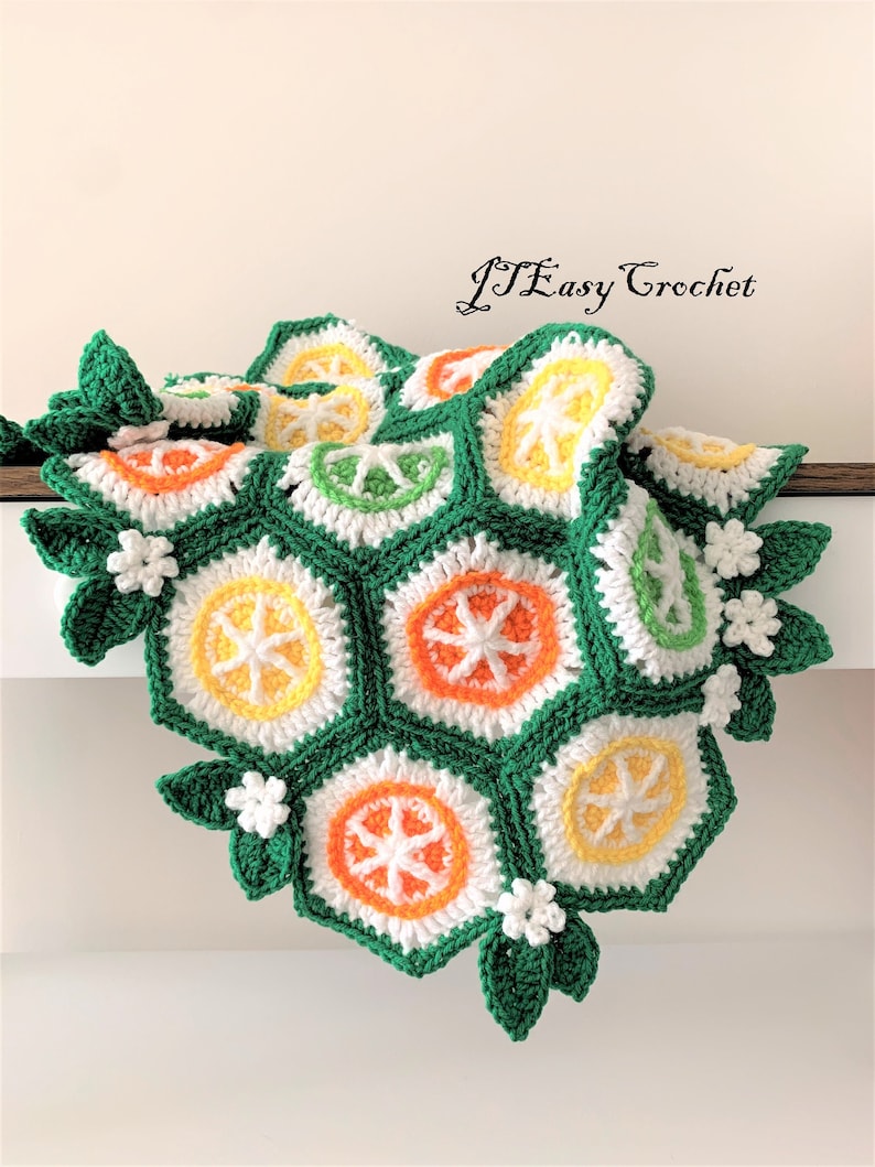 May include: A crocheted blanket with a citrus fruit design. The blanket is made of green, white, orange, and yellow yarn. The blanket is folded in half and has a white background.