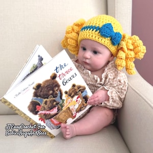 May include: A baby wearing a yellow crochet hat with blue accents and large yellow pigtails, holding a copy of "The Three Bears" book. The book cover features illustrations of bears and the title. The baby is sitting on a light-colored armchair.