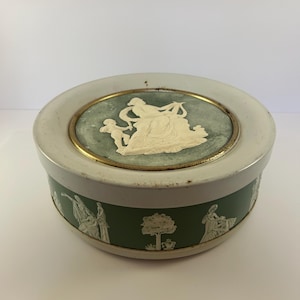 May include: Round, vintage trinket box with a white lid and a green base. The lid features a light green center with a white relief of figures. The base has white relief figures and a tree design. A gold-colored band encircles the lid.