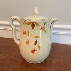 May include: Cream-colored ceramic pitcher with a lid, handle, and spout. The pitcher is decorated with brown and orange floral patterns and gold trim. The pitcher has vertical ribbed detailing and a pointed finial on the lid.