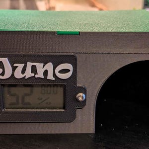 May include: A black and green terrarium with the name "Juno" on the front. It has a digital display showing humidity levels. The terrarium has an arched opening and a green top.